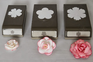 Drei blumenförmige Papierstanzer in unterschiedlichen Größen sind in einer Reihe angeordnet, jeweils gepaart mit einer wunderschönen Rose aus Papier davor: weiß, weiß-rosa und rosa.