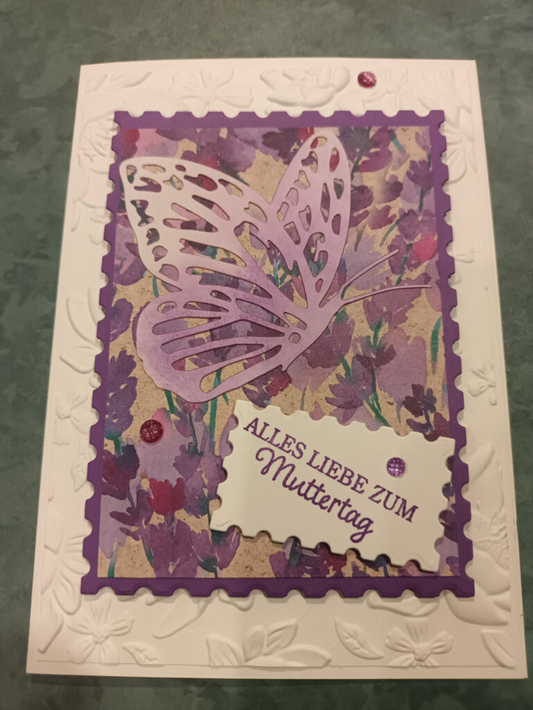 Eine Muttertagskarte mit einem dreidimensionalen lila Schmetterlingsdesign auf floralem Hintergrund, eingefasst von einem geprägten weißen Rahmen, mit dem Text „Mach was draus zum Muttertag.“