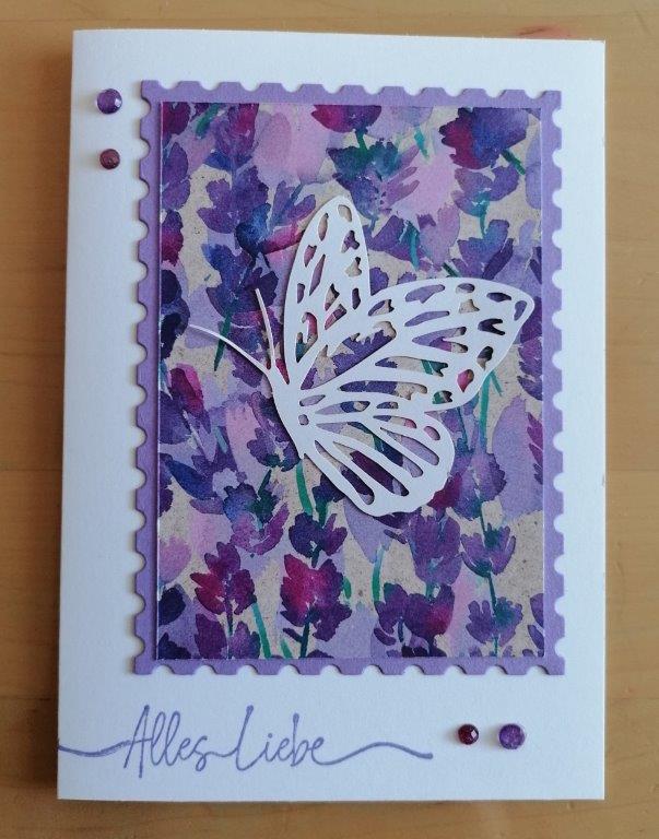 Handgefertigte Grußkarte mit Blumenmuster, einem weißen Schmetterlingsausschnitt und dem Satz „Mach was draus“ in Schreibschrift, verziert mit violetten Edelsteinen auf einer Holzoberfläche.