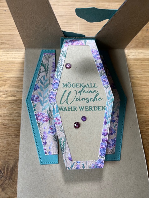 Eine dekorative Geschenkbox in Sargform mit floralem und blaugrünem Design, mit dem deutschen Satz „Mach was draus“ und zwei kleinen violetten Edelsteinen verziert.