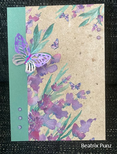 Handgefertigte Grußkarte mit lila Blumenaquarellmuster auf braunem Papier, geschmückt mit einem 3D-Schmetterling und Edelsteinverzierungen, signiert von Beatriz Punz für Mach was draus, 24. April.