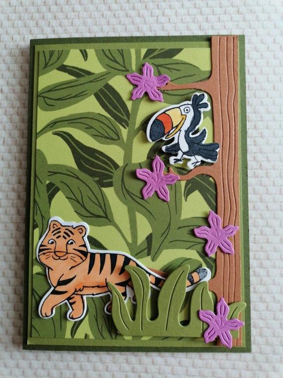 Eine handgemachte Karte mit ausgeschnittenen Verzierungen mit einem Tiger, einem Tukan auf einem Ast und rosa Blumen vor einem grünen Blätterhintergrund – perfekt für die Kreativ-Challenge „Mach was draus Mai 24“.
