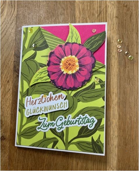Eine farbenfrohe Geburtstagskarte auf Holzoberfläche mit einer großen rosa Blume mit grünen Blättern und dem deutschen Text „Herzlichen Glückwunsch zum Geburtstag“. Die weitere Inschrift „Mach was draus Mai 24“ verleiht diesem lebendigen Gruß eine einzigartige Note.