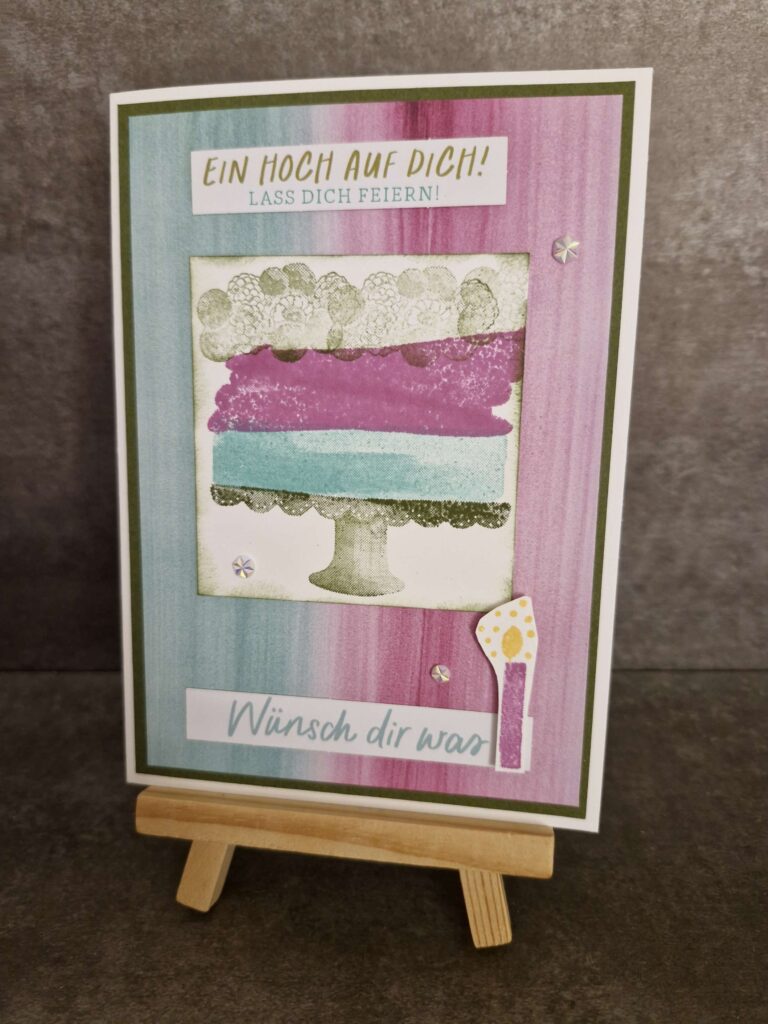 Grußkarte auf Holzständer mit farbenfrohem, abstraktem Kuchendesign, deutscher Text „Ein hoch auf dich! Lass dich feiern!“ und „Wünsch dir was“, übersetzt „Auf euch! Feiert!“ und „Wünsch dir was.“ Mach war am 24. August draus.