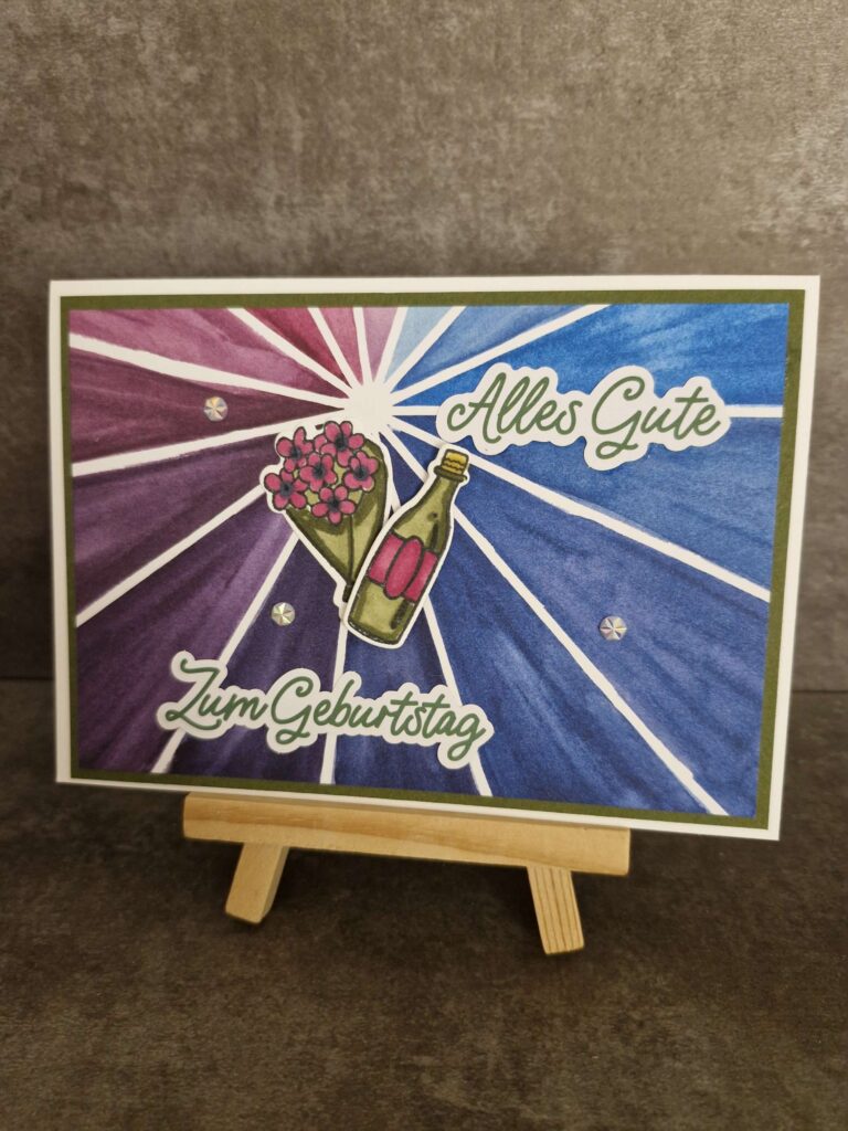 Eine Geburtstagskarte auf einer Staffelei trägt den deutschen Text „Alles Gute zum Geburtstag“ sowie Abbildungen eines Blumenstraußes und einer Flasche vor einem violett-blauen Sternenhintergrund. Mach was draus!