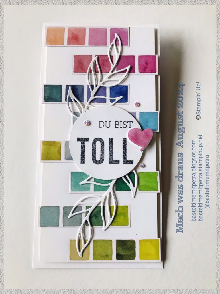 Handgemachte Karte mit dem deutschen Spruch "Du bist toll", umgeben von bunten Quadraten und einem Blattmuster. Die Karte ist mit kleinen Edelsteinen und einem rosa Herz verziert, perfekt für Mach was draus am 24. August. Text entlang des Randes.