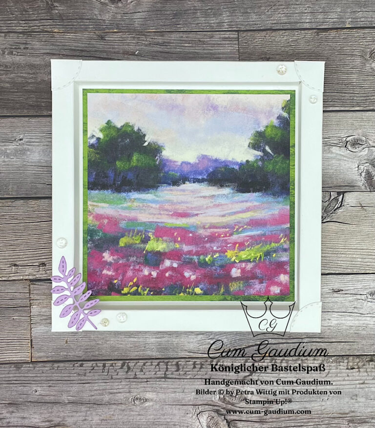 Ein gerahmtes Kunstwerk mit einer lebendigen Landschaft mit Bäumen, einer bunten Wiese und einem violetten Himmel. Der Rahmen steht auf einer Holzoberfläche und ist mit einem dekorativen violetten Blatt verziert. Perfekt für die Feier von Mach was draus am 24. September.