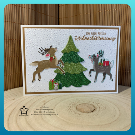 Eine festliche Weihnachtskarte mit zwei Rentieren, einem glitzernden grünen Baum und Geschenken auf einem strukturierten Hintergrund. Die Nachricht lautet: „Eine kleine Portion Weihnachtsstimmung? Mach was draus am 24. Oktober.“