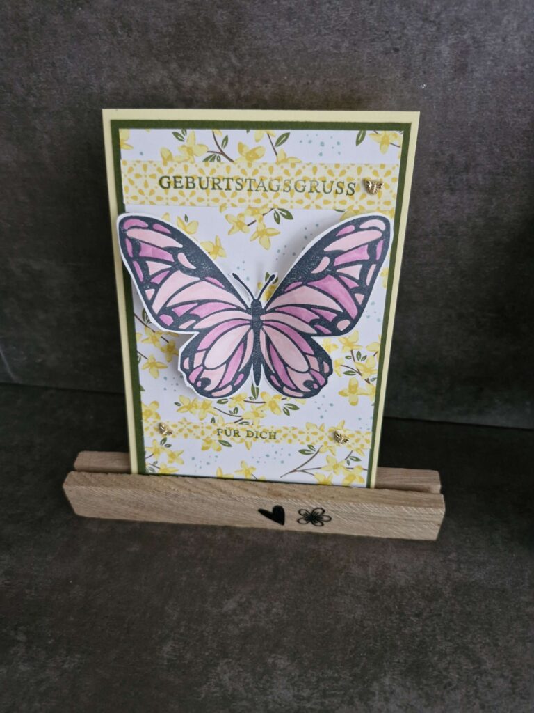 Grußkarte mit einem großen rosa Schmetterling und Blumenhintergrund, elegant auf einem Holzständer mit Herz- und Blumengravuren. Der Text lautet „Geburtstagsgruß für Dich“ und inspiriert Sie, den Tag zu genießen – Mach was draus, besonders am 25. Februar.