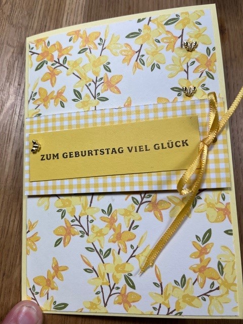 Gelb-weiße Geburtstagskarte mit Blumenmotiv und Schleife, auf der Vorderseite steht der deutsche Text „Zum Geburtstag viel Glück“. Perfekt, um jemandem am 25. Februar eine besondere Freude zu machen, also los, „Mach was draus“!.