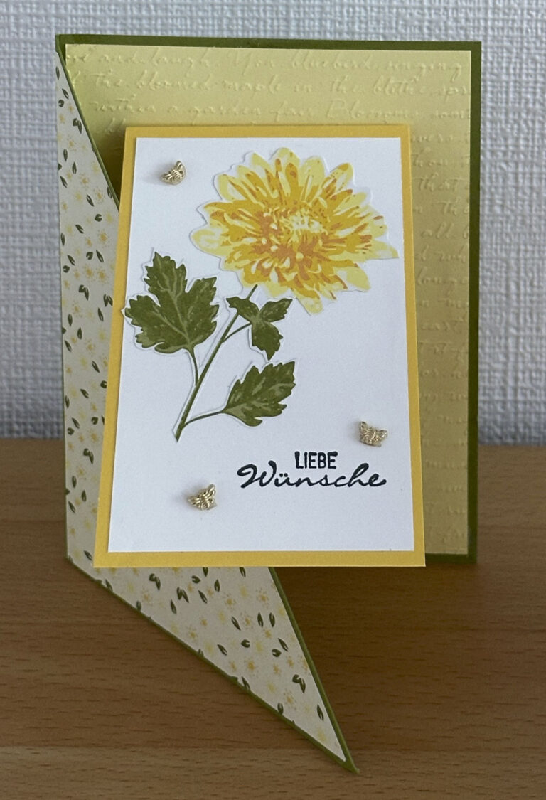 Grußkarte mit gelbem Blumenmuster und dem Text „Liebe Wünsche“, einem „Mach was draus 25. Februar“-Faltmuster und dekorativen Elementen auf einer Holzoberfläche.