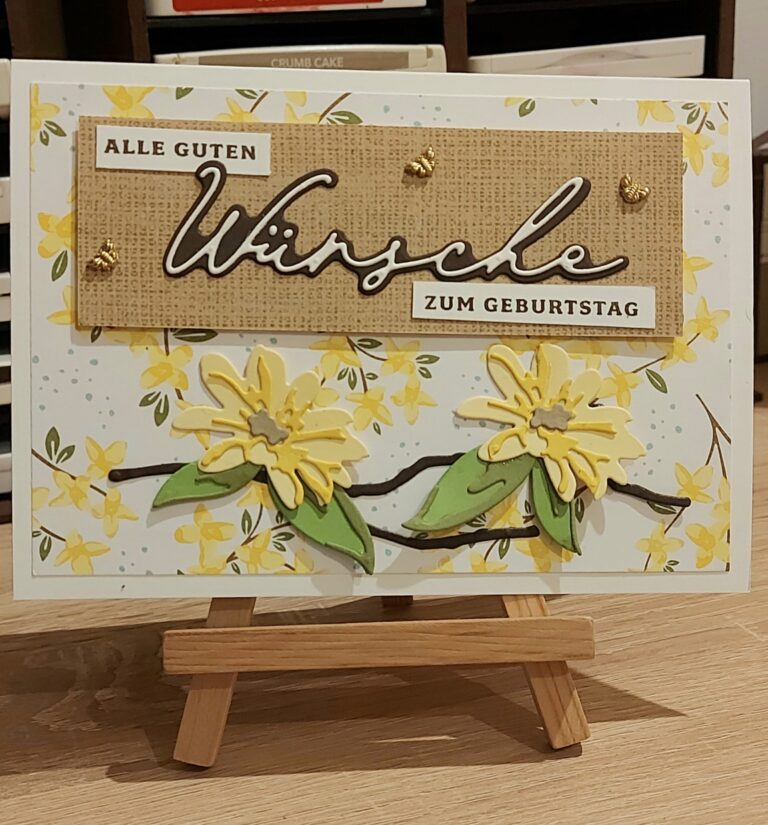 Handgefertigte Geburtstagskarte auf einer kleinen Holzstaffelei, mit gelben Blumen und dem deutschen Text „Alle guten Wünsche zum Geburtstag“ auf einem Blumenhintergrund. Ein perfektes Projekt für „Mach was draus“, bereit, am 25. Februar die Kreativität anzuregen.