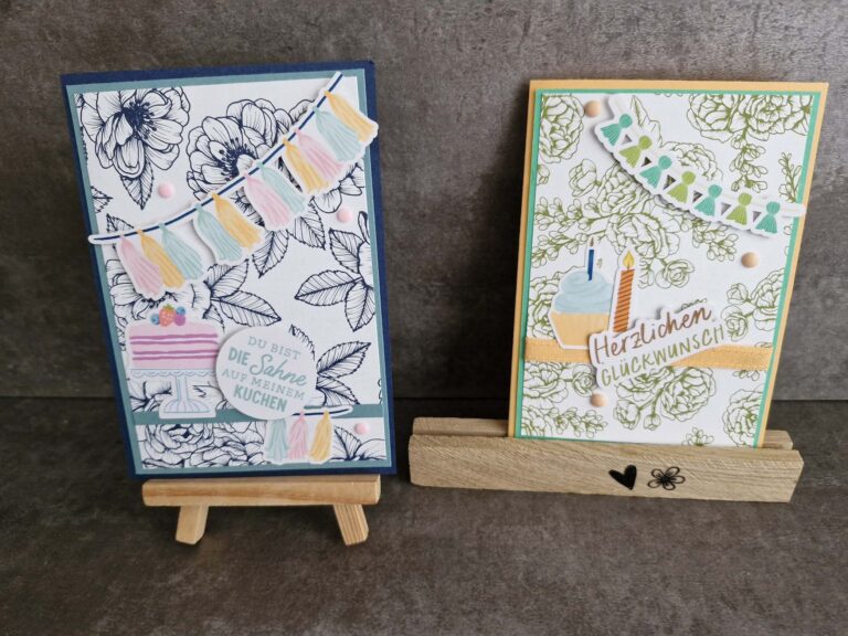 Zwei handgefertigte Geburtstagskarten mit floralen Hintergründen, Kuchen- und Bannerdekorationen aus der Mach was draus-Kollektion vom 25. April, präsentiert auf kleinen Holzständern vor einer grauen Oberfläche.