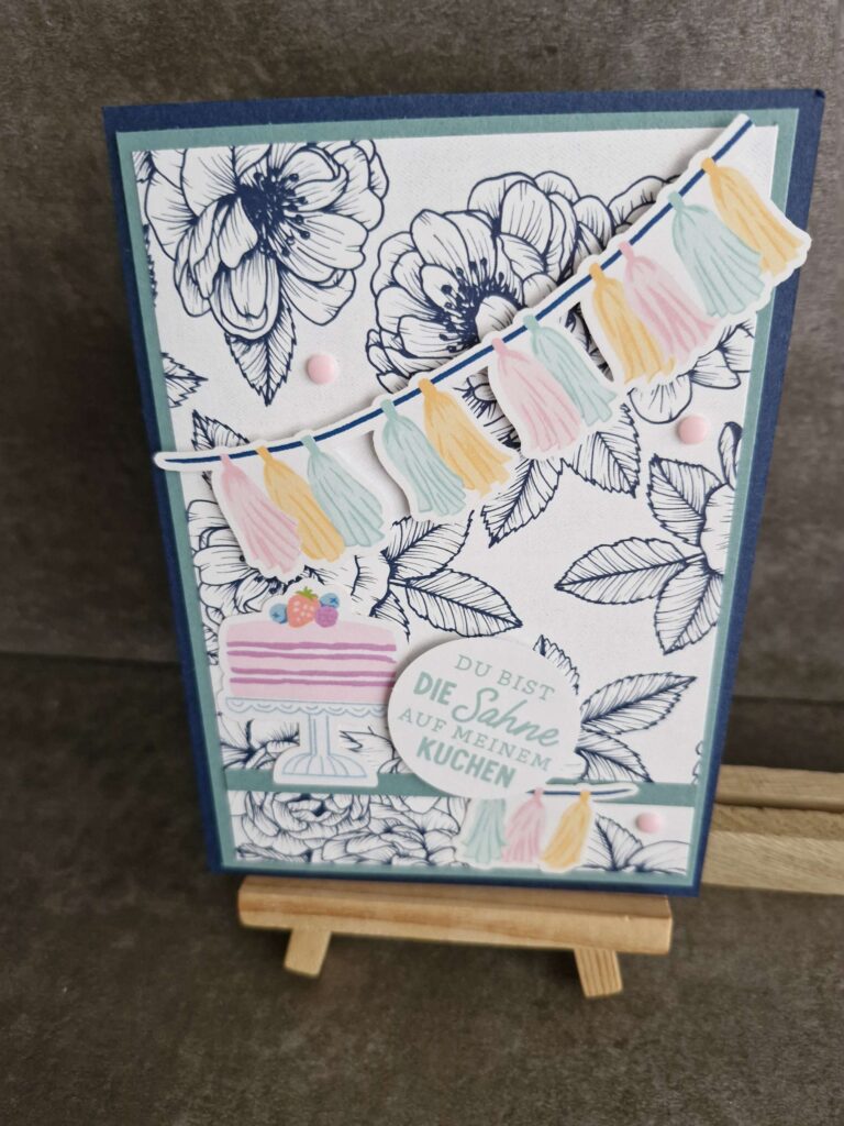 Eine handgemachte Grußkarte mit floraler Strichzeichnung, pastellfarbener Wimpelkette, einer Geburtstagstortenillustration und der deutschen Botschaft „Mach was draus, 25. April“, präsentiert auf einer kleinen Holzstaffelei.