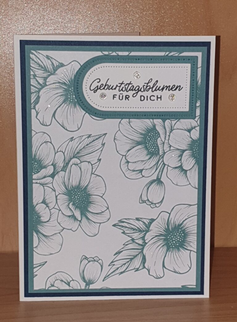 Eine Grußkarte mit blaugrünen Blumenillustrationen und einem Etikett mit der Aufschrift „Geburtstagsblumen für dich“ liegt auf einer Holzoberfläche – perfekt für Ihr Mach was draus-Projekt vom 25. April.