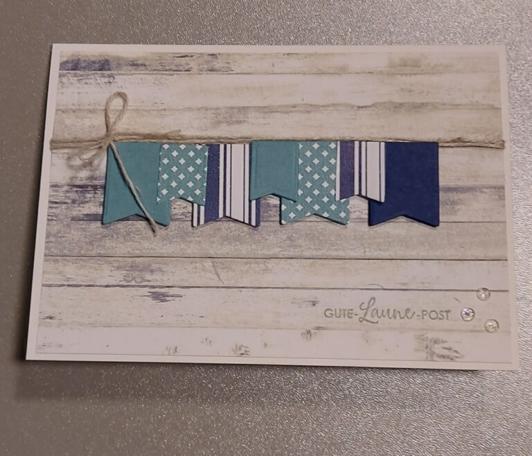 Eine handgemachte Karte von Mach was draus vom 25. April zeigt blaue und gemusterte Bannerausschnitte, die an einer Schnur auf einem Holzhintergrund hängen, mit dem Aufdruck „GUTE Laune-POST“ in der unteren rechten Ecke.