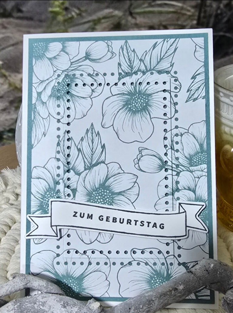 Eine Grußkarte mit blaugrünen Blumenillustrationen und einem Banner mit der Aufschrift „ZUM GEBURTSTAG“ liegt auf einer gewebten Oberfläche neben einer Kerze – die perfekte Inspiration für Ihr Mach was draus-Projekt vom 25. April.
