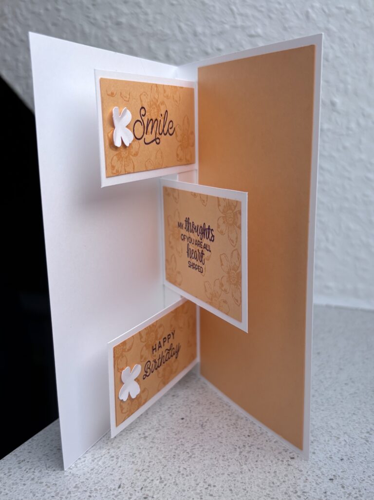 Eine handgemachte Geburtstagskarte, inspiriert von Mach war draus Mai 25, mit drei orangefarbenen Feldern im Inneren mit den Aufschriften „Lächeln“, „Gedanken an dich machen mein Herz glücklich“ und „Alles Gute zum Geburtstag“, alle mit zarten weißen Blumen verziert.