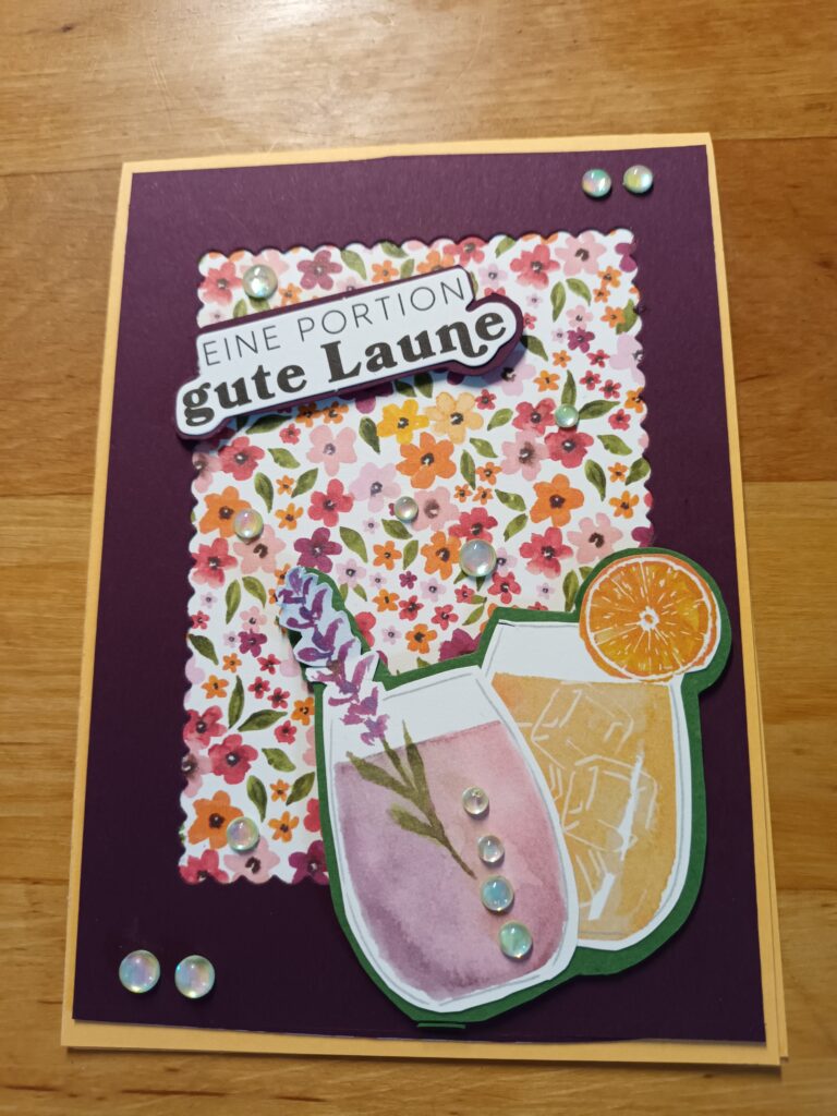 Eine handgemachte Karte von Mach was draus Mai 25 zeigt einen floralen Hintergrund, zwei illustrierte Getränke, Strassverzierungen und den deutschen Spruch „Eine Portion gute Laune“.