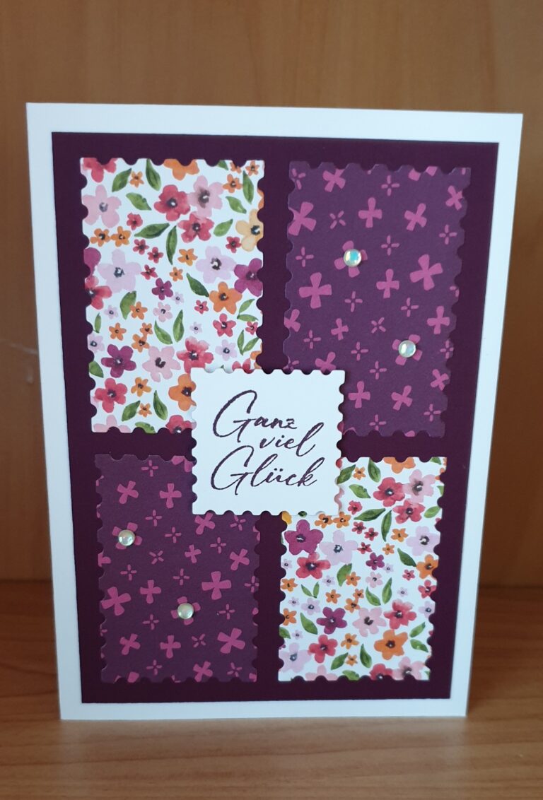 Eine handgemachte Grußkarte mit floralen und lila gemusterten Feldern, verziert mit kleinen Edelsteinen und einer zentralen Botschaft auf Deutsch, die lautet: „Ganz viel Glück“, inspiriert von „Mach was draus, 25. Mai“.