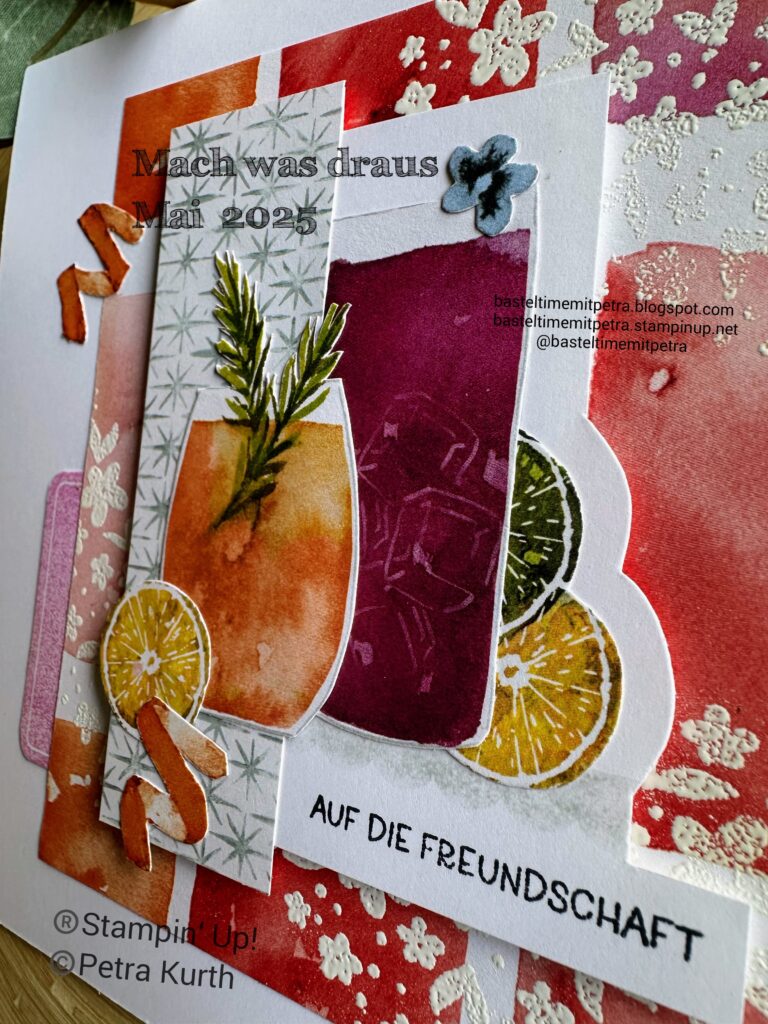 Eine handgefertigte Grußkarte mit einer farbenfrohen Getränkeillustration, geschnittenen Zitrusfrüchten, floralen Mustern und dem deutschen Text „Mach was draus Mai 25“. Die Karte enthält mehrschichtige Papierakzente und gestempelte Motive.