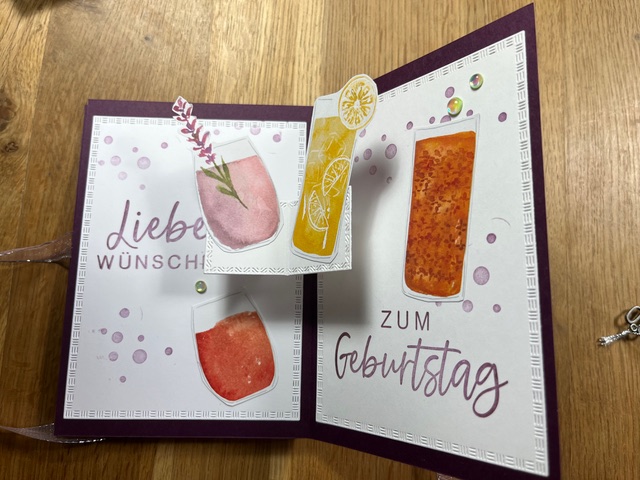 Eine handgefertigte Geburtstagskarte von Mach was draus Mai 25 zeigt farbenfrohe Getränkebilder, die deutschen Sätze „Liebe Wünsche“ und „Zum Geburtstag“ sowie dekorative Verzierungen, alles auf einer Holzoberfläche.