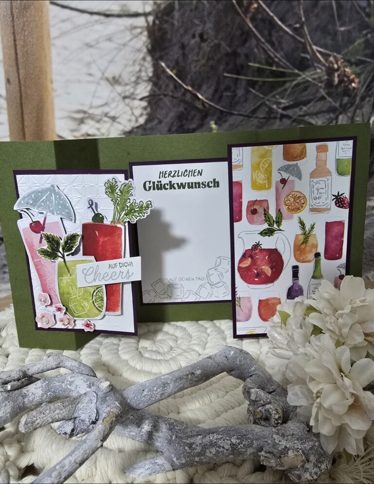 Eine handgemachte Geburtstagskarte mit farbenfrohen Cocktailillustrationen und deutschem Text von Mach was draus Mai 25, mit der Botschaft „Herzlichen Glückwunsch“, dargestellt auf einer strukturierten Oberfläche mit weißen Blumen und Zweigen.