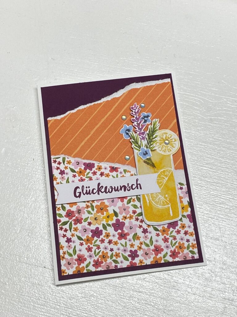 Handgemachte Grußkarte mit dem Text „Glückwunsch“, erstellt für Mach was draus Mai 25, mit floralen Mustern, einem orange gestreiften Hintergrund und einer Illustration eines Getränks mit Blumen und Zitronenscheiben.