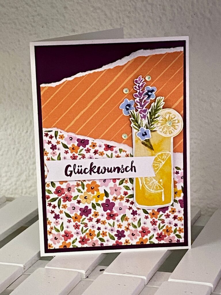 Eine handgemachte Grußkarte von Mach was draus Mai 25, mit dem Text „Glückwunsch“, einem Blumenmuster, orangefarbenen Streifen und einem Getränk mit Blumen und einer Zitronenscheibe auf der Vorderseite.