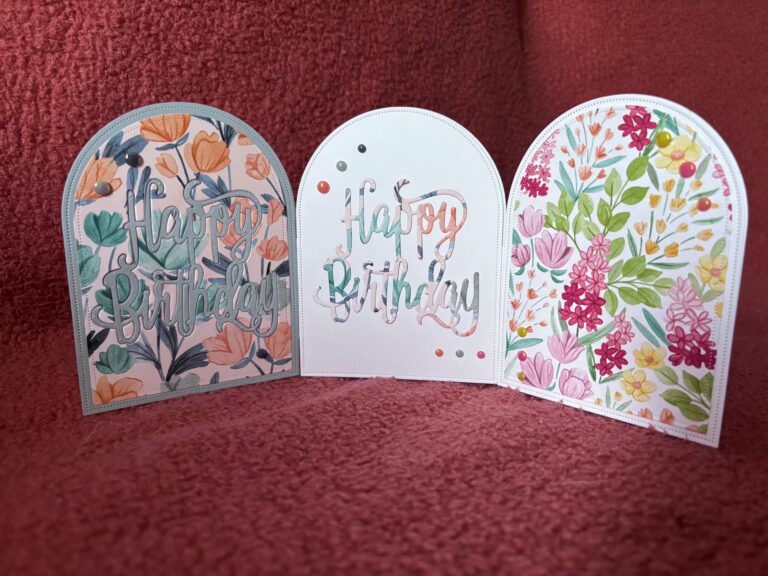 Drei gewölbte "Happy Birthday"-Karten mit floralen Motiven stehen aufrecht auf einer strukturierten rosafarbenen Oberfläche, die Teil der Kollektion Mach was draus Juni 25 ist.
