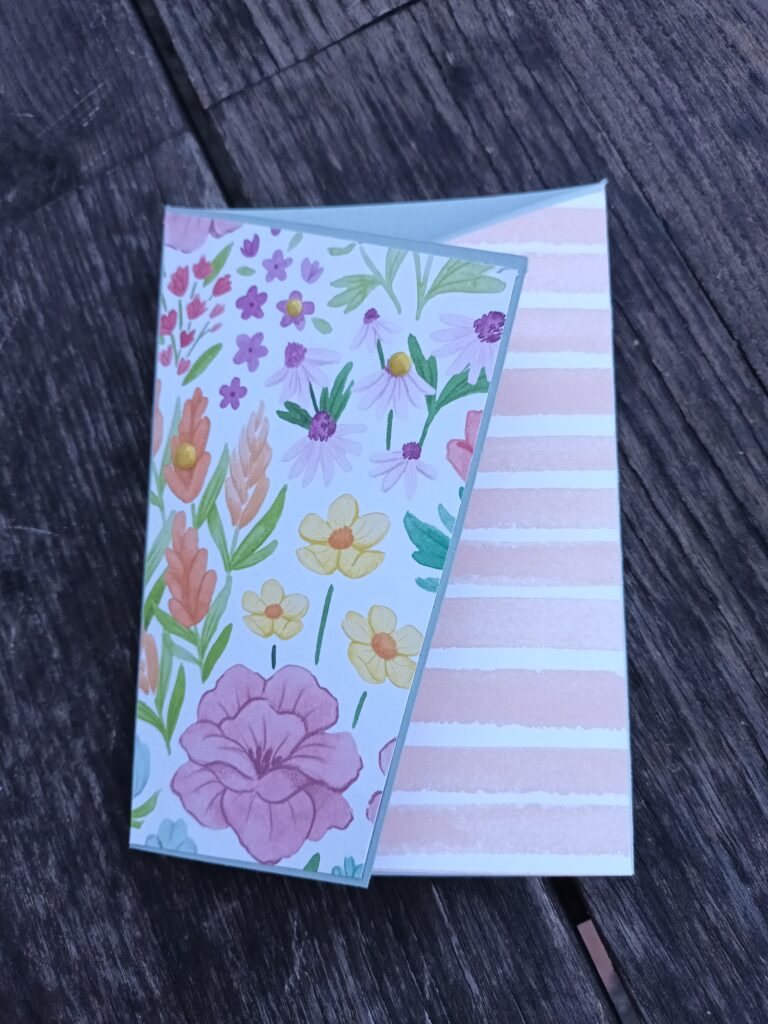 Eine Grußkarte mit geblümter Vorderseite und rosa gestreifter Innenklappe liegt auf einer Holzunterlage, die mit dem Spruch "Mach was draus Juni 25" für eine inspirierende Note sorgt.