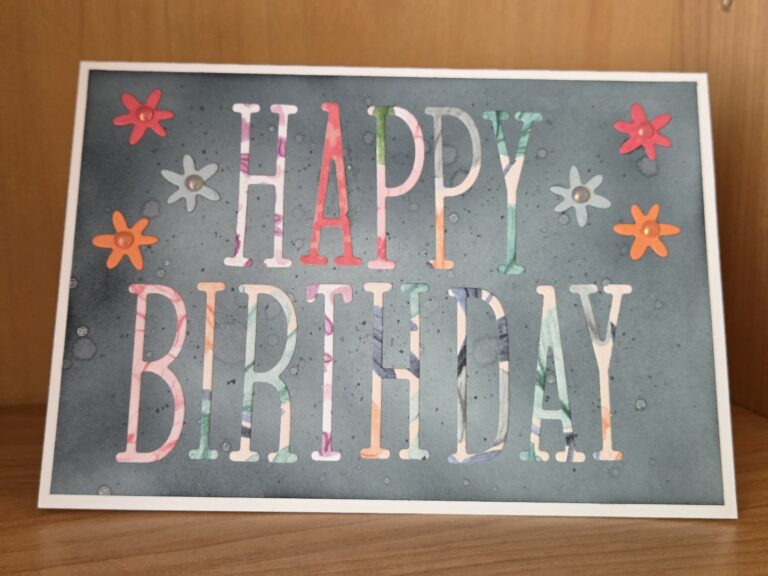 Eine Geburtstagskarte mit "HAPPY BIRTHDAY" in mehrfarbigen Buchstaben auf grauem Hintergrund, verziert mit orangefarbenen, rosa und weißen Blumenformen - perfekt zum Feiern von Mach was draus 25. Juni.