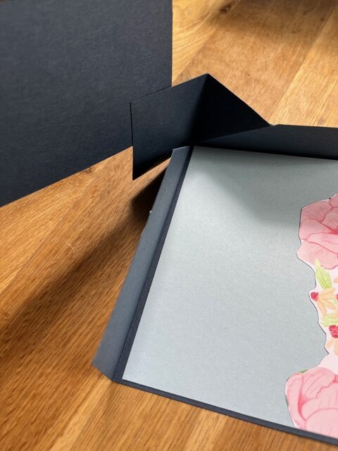 Ein offener schwarzer Ordner auf einer hölzernen Oberfläche enthält ein graues Blatt mit rosafarbenen Blumenmustern, die teilweise sichtbar sind und auf die Kreativität hinter Mach was draus 25. Juni hinweisen.