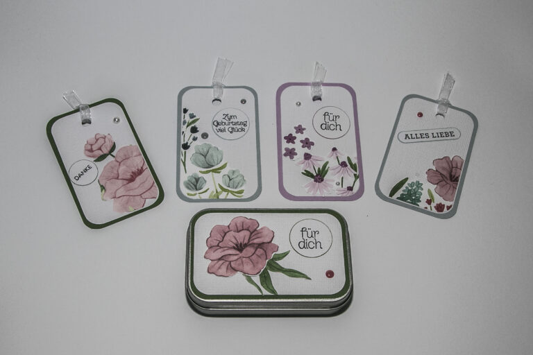 Fünf verzierte Geschenkanhänger mit floralen Motiven und deutschen Sprüchen aus Mach was draus Juni 25 sind auf weißem Hintergrund angeordnet, wobei ein Anhänger auf einer kleinen rechteckigen Dose platziert ist.