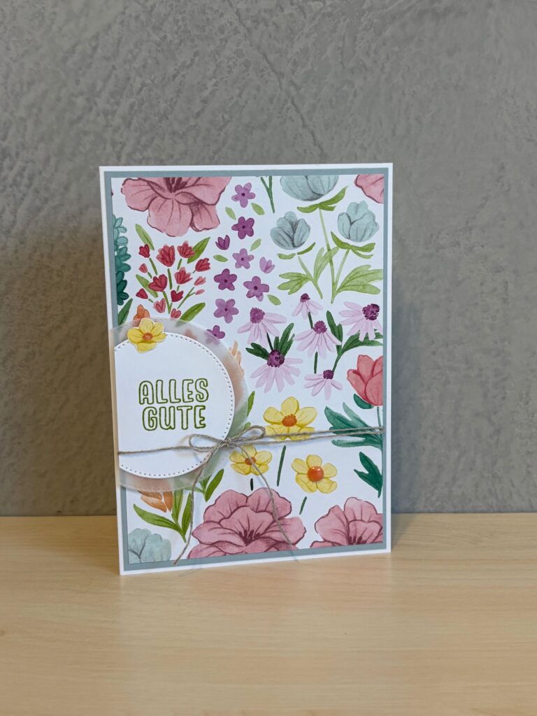 Eine Grußkarte mit einem farbenfrohen Blumenmuster und dem deutschen Satz "Alles Gute" auf einem weißen Etikett, perfekt für Mach was draus 25. Juni, steht auf einer hellen Holzfläche vor einer grau strukturierten Wand.