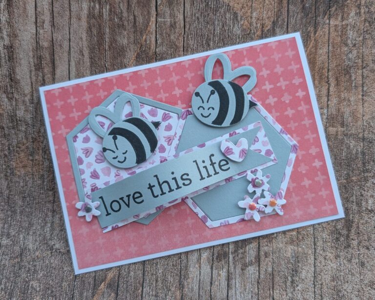 Eine handgefertigte Karte für Mach was draus 25. Juni ruht auf einer Holzoberfläche und zeigt zwei Cartoon-Bienen, rosa Blumenformen und den Text "love this life" auf einem grauen Banner.
