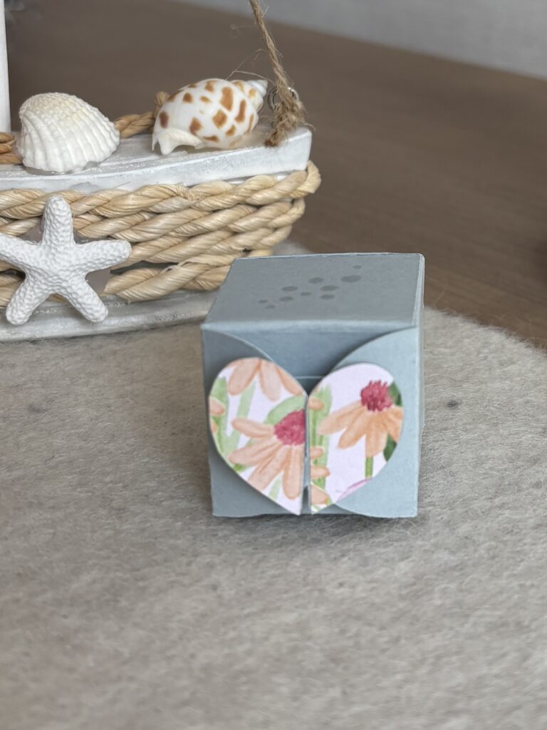 Eine kleine blaue Papierschachtel mit einem floralen Herzmotiv auf der Vorderseite, Teil der Kollektion Mach was draus Juni 25, steht auf einer hellen Fläche mit einem Korb mit Muscheln und einem Seestern im Hintergrund.
