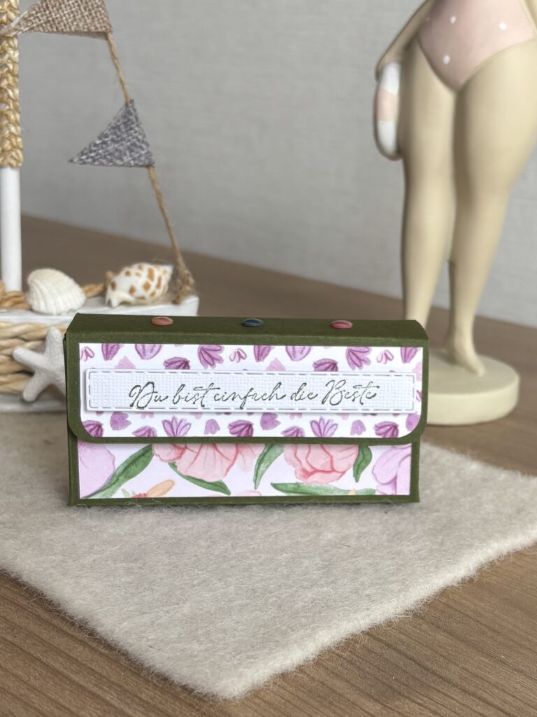 Eine dekorative Schachtel mit einem Blumenmuster und dem deutschen Spruch "Du bist einfach die Beste" steht auf einem Tisch mit maritimem Dekor im Hintergrund, inspiriert von Mach was draus Juni 25.