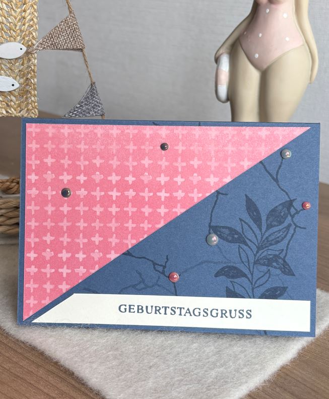 Eine blaue und rosafarbene handgemachte Geburtstagskarte von Mach was draus 25. Juni zeigt den deutschen Text "GEBURTSTAGSGRUSS" und Blattmuster, verziert mit kleinen Perlen. Eine dekorative Figur und ein Ornament befinden sich im Hintergrund.
