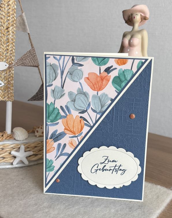 Eine handgefertigte Geburtstagskarte mit einem floralen und strukturierten blauen Design, die für Mach was draus 25. Juni erstellt wurde, steht aufrecht auf einem Tisch neben einem dekorativen maritimen Ornament und einer Keramikfigur.