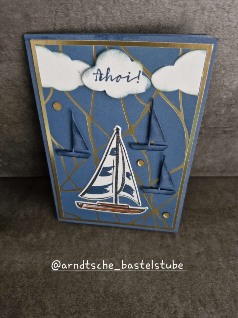 Handgemachte nautische Grußkarte mit Segelboot, gefalteten Papierbooten, goldenen Linien und "Ahoi!" auf weißen Wolkenformen. Erstellt für Mach was draus 25. Juli. Instagram-Handle @arndtsche_bastelstube am unteren Rand.