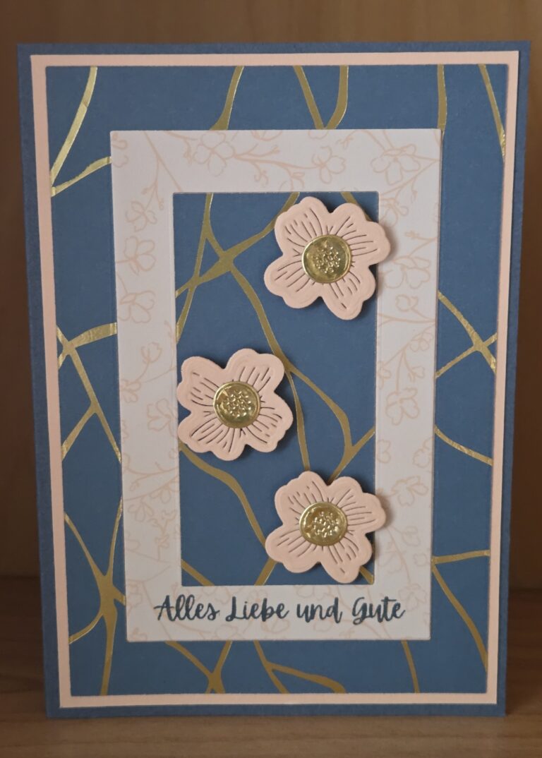 Eine handgefertigte Grußkarte, inspiriert von Mach was draus 25. Juli, zeigt einen blau-gold gemusterten Hintergrund, einen weißen Blumenrahmen und drei rosa Blumen mit goldenen Zentren. Auf der Unterseite steht "Alles Liebe und Gute.