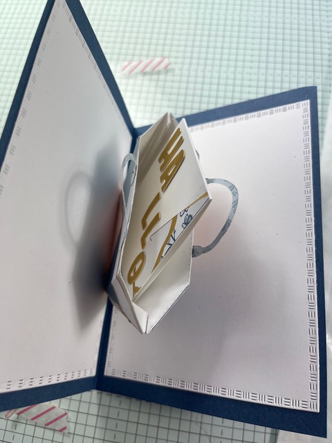 Eine handgefertigte Pop-up-Grußkarte zeigt eine dreidimensionale Papiertüte mit dem Schriftzug "HELLO" auf einem blau-weiß gemusterten Hintergrund - die perfekte Inspiration für Mach was draus 25. Juli.
