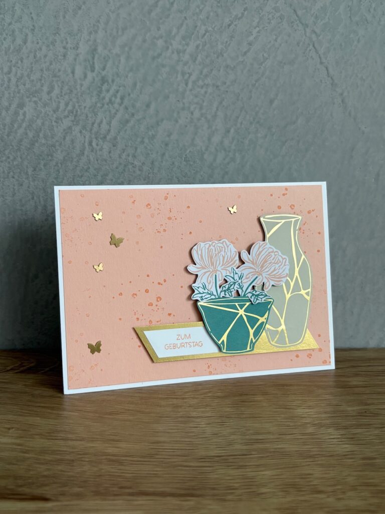 Grußkarte mit einer Vase und Topfblumen auf einem pfirsichfarbenen Hintergrund, goldenen Akzenten, kleinen Schmetterlingsausschnitten und einem Schild mit der Aufschrift "Zum Geburtstag" - inspiriert von Mach was draus 25. Juli.