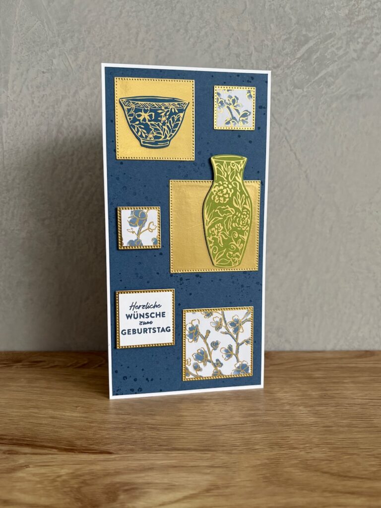 Eine handgefertigte Geburtstagskarte mit blauem und goldenem Design, auf der eine Schale, eine Vase und Blumenmuster abgebildet sind, steht aufrecht auf einer hölzernen Oberfläche - perfekt, um Mach was draus 25. Juli stilvoll zu feiern.