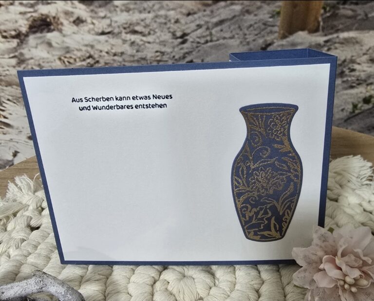Eine Grußkarte mit einer blau-goldenen Vasenillustration und dem deutschen Text "Aus Scherben kann etwas Neues und Wunderbares entstehen" auf einer strukturierten Oberfläche - perfekt für die Inspiration von Mach was draus 25. Juli.