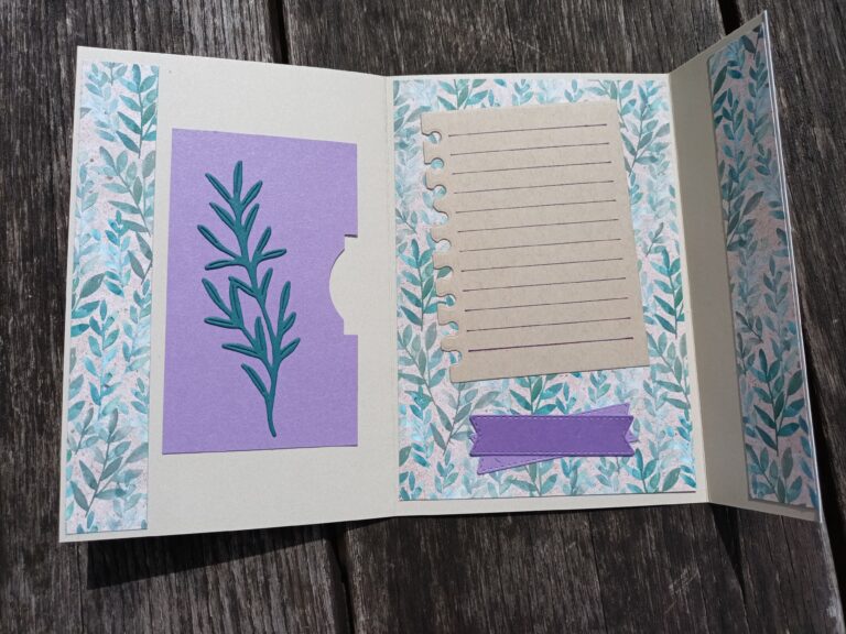 Eine handgefertigte Karte für "Mach was draus 25. August" hat eine blumengemusterte Umrandung, eine violette Tafel mit einem Blattausschnitt auf der linken Seite und gefüttertes Notizpapier mit einem violetten Band auf der rechten Seite, das offen auf Holz ausgestellt ist.