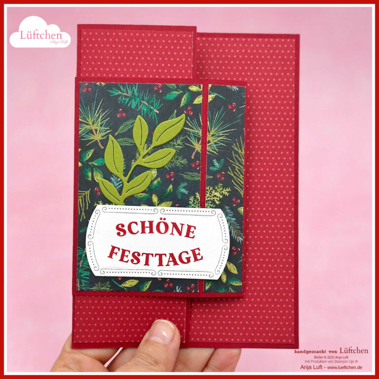 Eine Hand hält eine rote, gemusterte Festtagskarte mit grünem und goldenem Blattwerk, die perfekt zum Neuen Mini 25 passt, und ein Etikett mit der Aufschrift "Schöne Festtage".