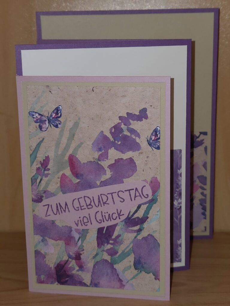 Eine handgefertigte Geburtstagskarte mit Blumen- und Schmetterlingsmotiven, die auf der Vorderseite den deutschen Text "Zum Geburtstag viel Glück" trägt. Inspiriert von Mach was draus 25. August, sind zwei ähnliche Karten dahinter platziert.