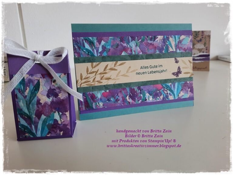 Eine handgefertigte Karte mit Blumenmustern, dem deutschen Text "Alles Gute im neuen Lebensjahr!" und einer passenden lilafarbenen Geschenkbox von Mach wurde am 25. August elegant auf einer weißen Fläche draus.